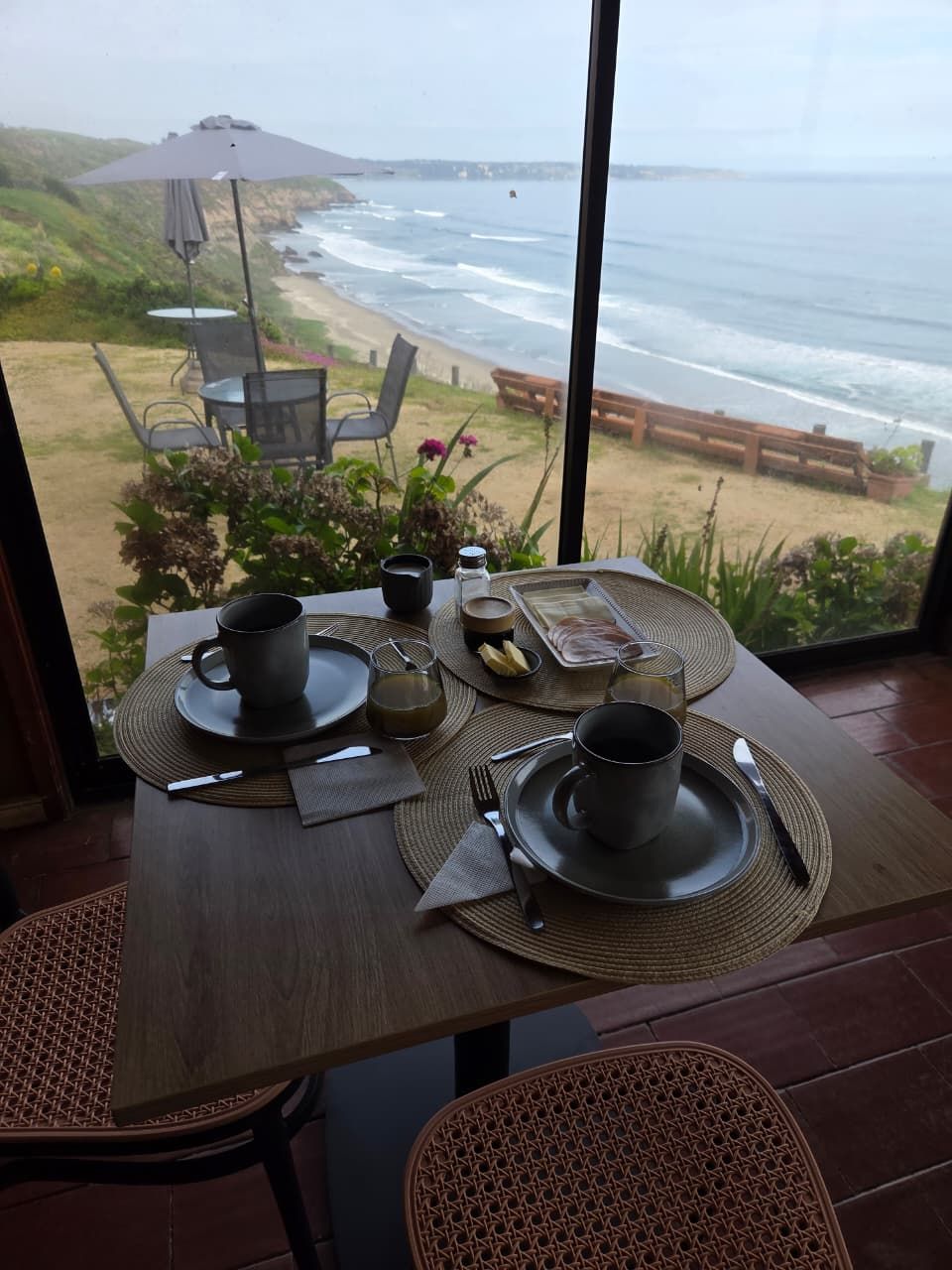 Desayuno servido en una mesa junto a la ventana, con el jardín y el mar abajo