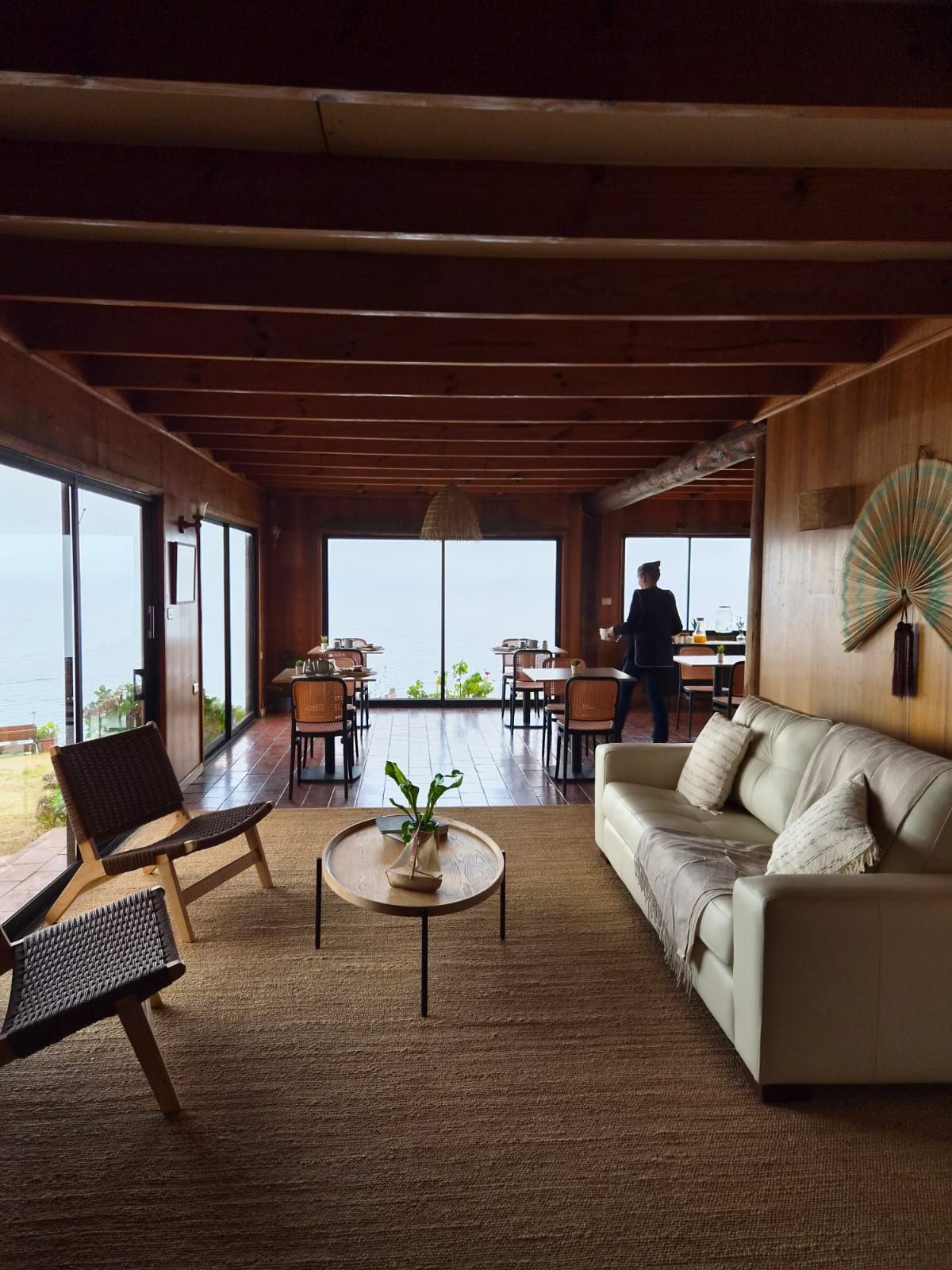 Living de la casona con sofá, sillas de cuerda y vigas de madera, con el comedor y los ventanales al fondo